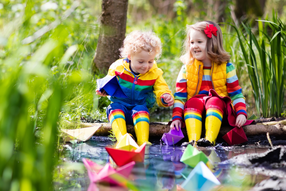 Children in brightly colored clothes playing in puddles with paper boats - Rainy Day Activities for Families - Kids Outside Adventures