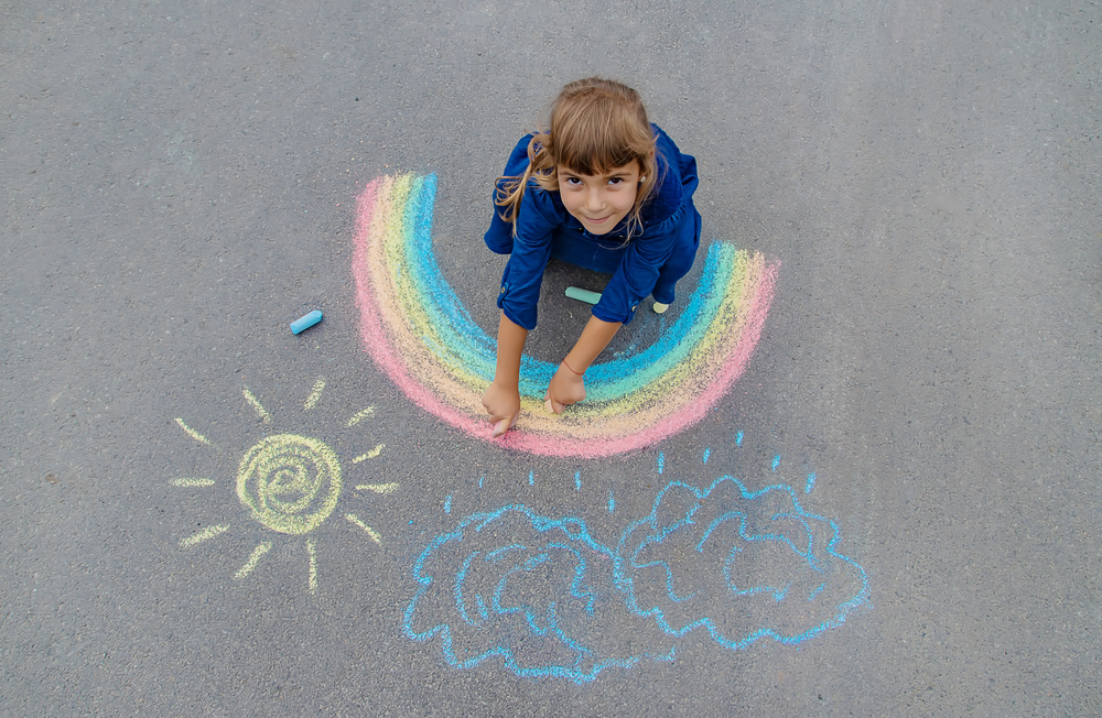 Child drawing with sidewalk chalk on the pavement pictures of a sun, rain clouds, and a rainbow - Rainy Day Activities for Families - Kids Outside Adventures