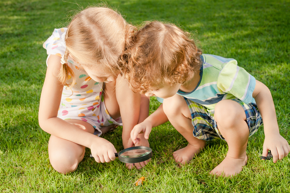 Two young children looking at bugs in the grass through a magnifying glass - Teaching Kids to Appreciate Nature - Kids Outside Adventures