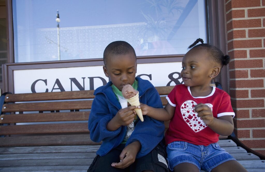 Two children sitting on a bench outside a store sharing an ice cream cone - Kids Outside Adventures